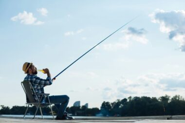 The image depicts a beginner fisherman sitting comfortably in a chair while fishing, enjoying a drink under a clear sky. The serene setting, with the fishing rod extended and the backdrop of a distant cityscape and lush greenery, captures the essence of relaxation and the simple pleasures of fishing.