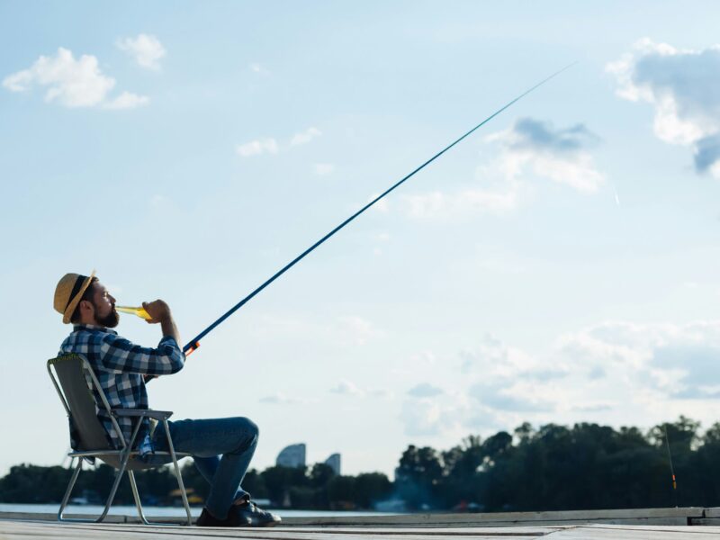 The image depicts a beginner fisherman sitting comfortably in a chair while fishing, enjoying a drink under a clear sky. The serene setting, with the fishing rod extended and the backdrop of a distant cityscape and lush greenery, captures the essence of relaxation and the simple pleasures of fishing.